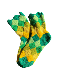 The image shows a pair of hand-crocheted socks with a pattern made up of small diamond-shaped patches. The colors alternate in bright greens, yellows, and yellow-oranges, creating a diagonal patchwork effect. The socks are partially overlapping, with the 