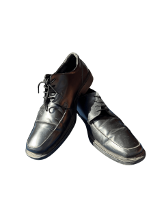 The image shows a pair of black men’s leather dress shoes. The shoes have low heels, laces, and visible stitching on the toe area. One shoe is upright, the other leans against it sideways.