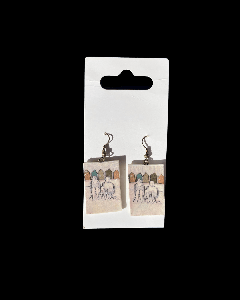 The image shows a pair of small dangling earrings attached to a white card. Each earring features a rectangular pendant with a delicate illustration of two people sitting on a beach, with colorful little houses in the background. The artwork is drawn with
